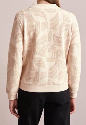 Texturiertes Creme-Sweatshirt mit einem Blättermuster, gerippten Bündchen und Saum sowie einem runden Halsausschnitt, kombiniert mit schwarzen Hosen.