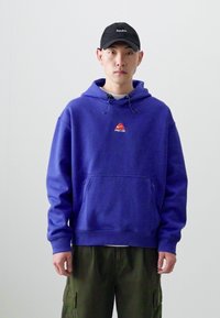Felpa viola con tasca frontale, cappuccio regolabile con cordino e un piccolo logo rosso. Indossata con pantaloni cargo verde oliva. Cappellino nero.