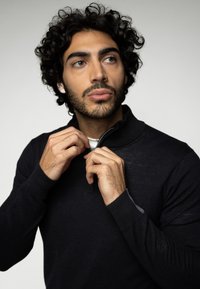 Pulover negru cu fermoar, cu manșete ribbate; modelul ajustează gulerul. Materialul pare moale, cu o textură netedă. Accente vizibile pe mâneci.
