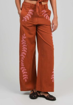 Pantalon ample de couleur rouille avec des motifs de feuilles brodées roses, porté avec des sandales tissées noires, mains reposant près des poches.