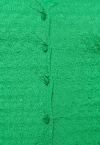 Zelená texturovaná látková blůza s vlnitým vzorem, se pěti odpovídajícími knoflíky. Materiál vypadá lehce a je tkaný.