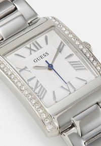 Ceas rectangular argintiu cu un cadran texturat, cifre romane, ace albastre și accente de cristal pe bezel. Numele brandului este afisat proeminent.