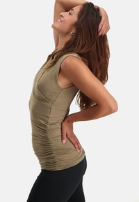 Olivgrön ärmlös topp med rynkad sidodetalj, kombinerad med svarta leggings. Tyget är slätt med en figurnära silhuett.