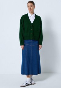 Vrouw in een donkergroen knoopcardigan over een witte blouse, blauwe denim midi rok, witte sokken en witte sneakers met zwarte strepen.