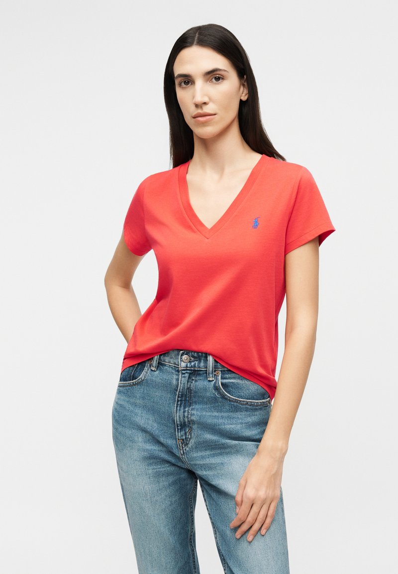 Vrouw met lang, donker haar, gekleed in een rood T-shirt met V-hals en blauwe jeans, staand met één hand in haar zak tegen een effen achtergrond.