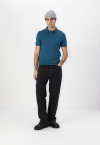 Teal polo skjorte med krage, kombinert med løse svarte jeans og svarte sko. Personen har på seg en lys grå strikket lue. Ensfarget bakgrunn.