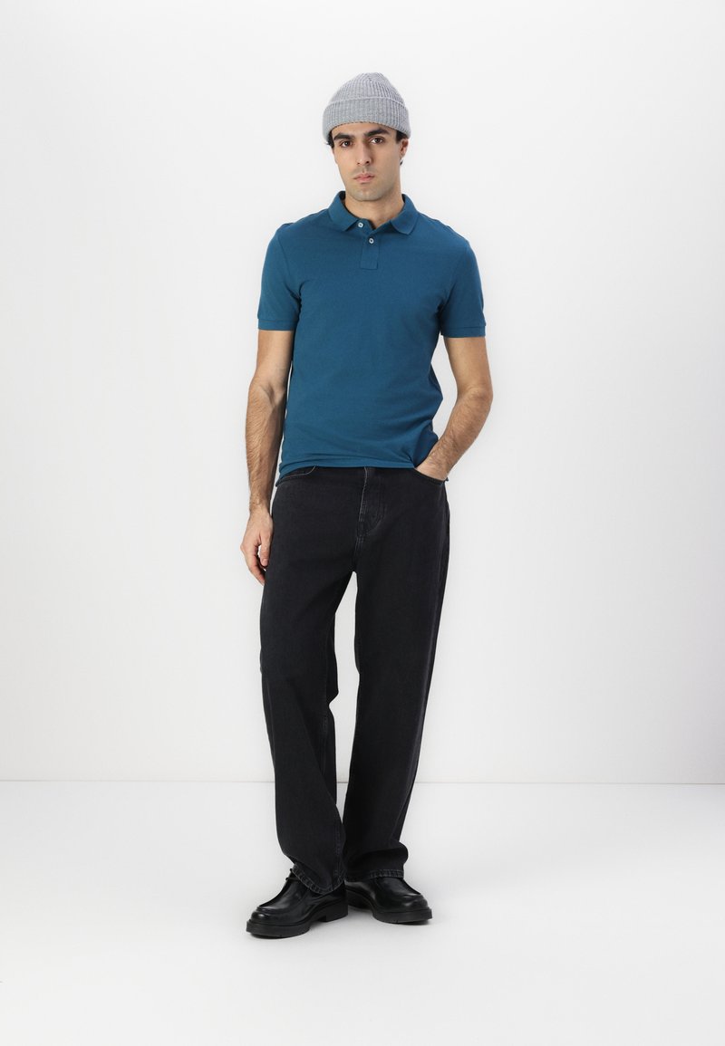 Teal polo skjorte med krage, kombinert med løse svarte jeans og svarte sko. Personen har på seg en lys grå strikket lue. Ensfarget bakgrunn.