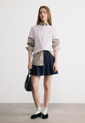 Jonge vrouw met een gelaagd hemd en een roze T-shirt, denim rok, witte sokken met zwarte platte schoenen, en een zwarte gevlochten handtas in haar hand, staand tegen een effen muur.