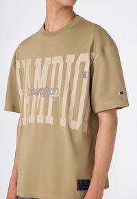 Persona vistiendo una camiseta beige de Champion de gran tamaño con grandes textos y logotipos en blanco y gris, cortada a la altura de los hombros y la cintura.