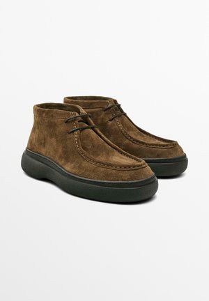 Stivali ankle in suede marrone con suole in gomma nera e due occhielli per lacci neri, mostrati a coppia su uno sfondo bianco.
