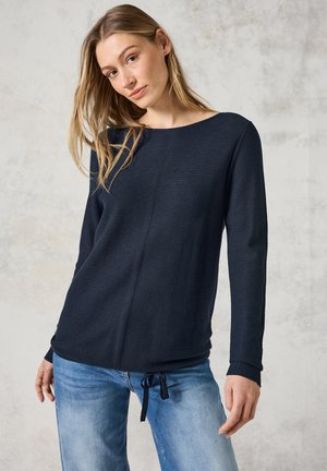 Top in maglia blu navy con texture, scollatura ampia, maniche lunghe e vestibilità dritta. Presenta un dettaglio a fiocco sul fondo. Indossato con jeans blu chiaro.