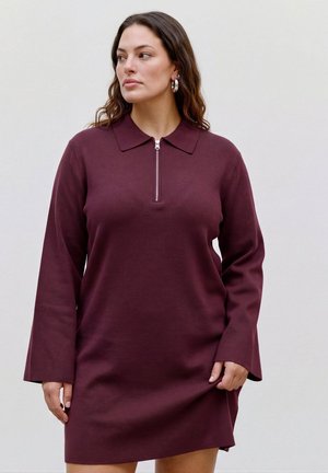 Femme aux longs cheveux portant une robe bordeaux zippée avec un col et des manches longues, debout devant un fond clair uni.