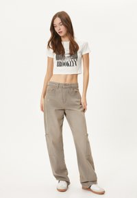 Kraftigt hvidt t-shirt med sort grafisk tryk, parret med løse khaki cargo-bukser med lommer og subtil slidtegn. Hvide sneakers.