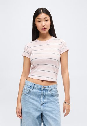 Jonge vrouw met stijl zwart haar, die een lichtroze gestreept cropped t-shirt en een lichtblauwe high-waisted jeans draagt, staand stil.
