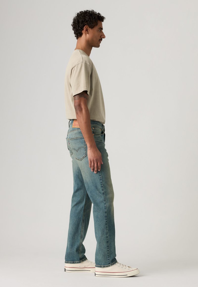 Mand i beige T-shirt, blå jeans og hvide sneakers stående sidelæns mod en ensfarvet lys baggrund.