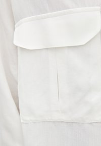 Tissu blanc à la texture lisse avec une poche avant et un pli vertical sous la poche. Les bords sont soigneusement cousus.