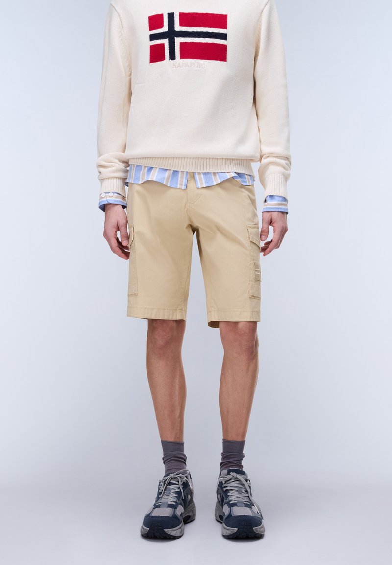 Hombre con pantalones cortos cargo beige, suéter crema con diseño de bandera roja y azul, camisa a rayas debajo, calcetines grises y zapatillas deportivas negras.