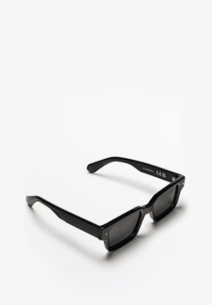 Lunettes de soleil noires avec une monture carrée, fabriquées en plastique brillant. Verres sombres, design simple, avec des branches angulées et un détail de logo sur le côté.