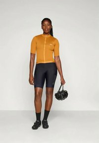 Femme portant un maillot de cyclisme moutarde et un short de cyclisme noir, tenant un casque de vélo noir dans la main droite, se tenant contre un fond uni.