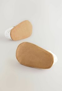 Vita tofflor med en strukturerad ovansida och en mjuk brun mockasula. Rundad form med sydda detaljer längs kanterna.