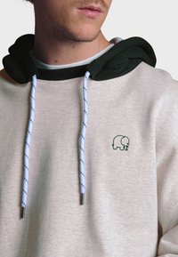 Sudadera color beige claro con capucha verde oscuro, con cordones azules con puntos negros y un pequeño logo de un elefante verde en el pecho.