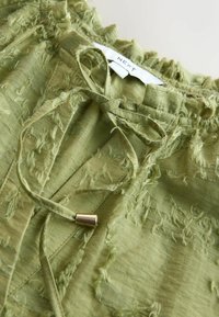 Jasnozielona bluzka z teksturowanej tkaniny z detalami w postaci postrzępionych akcentów, wiązaniem przy dekolcie oraz małym metalowym końcówką sznurka, marki Next.