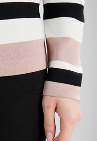 Manica di un maglione a righe con ampie bande orizzontali nere, bianche e beige, che termina sopra una mano con unghie naturali contro pantaloni neri.
