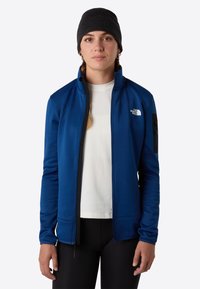 Modrá fleece bunda s vysokým límcem, zapínáním na zip a černou kapsou jako akcentem. Nosená přes bílé tričko, doplněná o černou čepici.