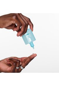 Lys blå klemflaske med en spiss tupp, som dispenserer en liten dråpe klar væske i en utstrakt hånd. Glatt tekstur og enkelt design.