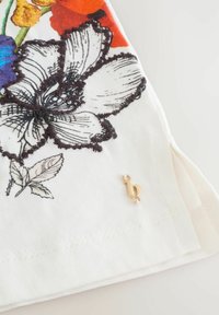 Witte stof met kleurrijke bloemenborduurwerk in rood, blauw en zwart. Bevat een klein accent van gouden hardware vlakbij de zoom.