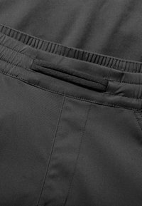 Schwarzes Stoffkleidungsstück mit strukturierter Oberfläche, elastischem Bund und einer dezenten Gesäßtasche mit glatter Oberfläche.