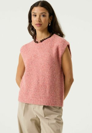 Vrouw met lang donker haar draagt een roze mouwloos gebreid topje, beige broek, bruine kralenketting en gouden oorringen, staand tegen een witte achtergrond.