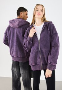 Felpa con zip viola, dalla vestibilità ampia, con tasche anteriori e cappuccio. Il tessuto ha un aspetto sbiadito e testurizzato. Modelli che indossano abbigliamento casual.