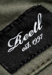 Schwarz gewebtes Etikett mit aufgesticktem weißen Text "Reell" und "est. 1997", angenäht auf einem grünen Textilhintergrund.