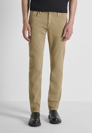 Hombre vestido con pantalones beiges ajustados y botas de cuero negras de pie contra un fondo claro y liso.