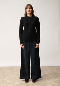 Czarny prążkowany sweter z dzianiny zestawiony z szerokimi spodniami z ciemnego sztruksu. Modelka stoi w neutralnym otoczeniu, ukazując teksturę i fason stroju.