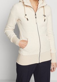 Sudadera con cremallera en color beige claro, fabricada en un material suave. Cuenta con una capucha, cordones, bolsillos frontales y puños acanalados. Textura lisa con un patrón sutil.