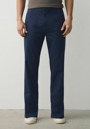 Mann trägt marineblaue gerade geschnittene Hose und weiße Sneakers und steht drinnen vor einer einfachen Wand.