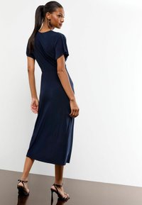 Navyblaues Midikleid mit kurzen Ärmeln, gerafftem Rückendetail und glatter Stofftextur. Kombiniert mit schwarzen High-Heel-Sandalen.