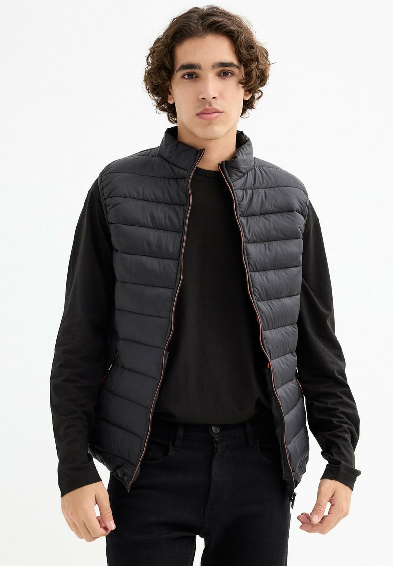 Chaleco de plumas negro con cuello alto, con costuras horizontales, cremallera frontal y bolsillos laterales, llevado sobre una camiseta de manga larga negra.