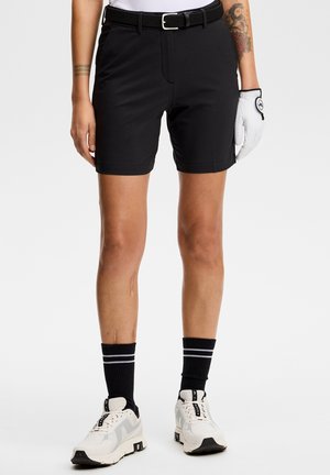 Person iført sorte shorts med sort bælte, hvid golfhandske på højre hånd, sorte sokker med hvide striber og hvide sneakers.