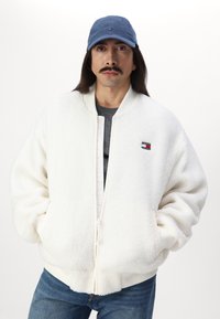 Chaqueta bomber de felpa blanca con cremallera en la parte delantera, puños acanalados y un pequeño parche con el logo. Combinada con una gorra azul clara y una camiseta gris.