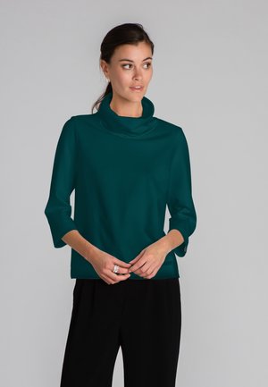 Tyrkysový svetr s vykrojeným límcem a 3/4 rukávy, vyrobený z hladkého materiálu. Design se vyznačuje uvolněným střihem a jemnou texturou, v kombinaci s černými kalhotami.