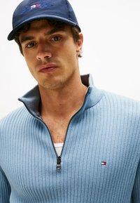 Lichtblauwe geribde zip-up trui met hoge kraag, voorzien van een klein Tommy Hilfiger-logo en een marineblauwe pet met geborduurde branding.
