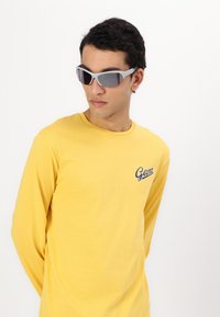 Gelbes Langarmshirt mit Rundhalsausschnitt, ausgestattet mit einem blauen Logo. Das Modell trägt graue Sonnenbrillen mit dunklen Gläsern und einer enganliegenden Passform.