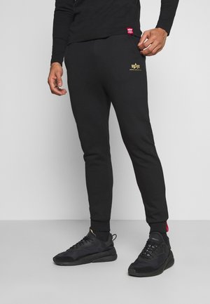 Mężczyzna w czarnych spodniach dresowych z logo Alpha Industries i czarnych sneakersach, stojący na szarym tle.