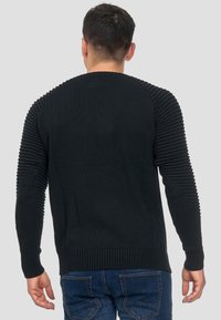 Pull en tricot noir avec texture côtelée sur les épaules et les manches, présentant un ourlet arrondi et des manches ajustées. Vue de dos affichée.