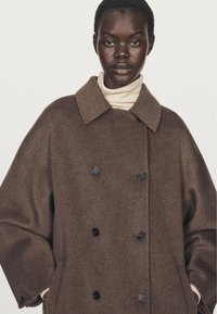 Brun dubbelknäppt kappa i mjuk ull med bred krage och svarta knappar. Har en avslappnad passform och långa ärmar.