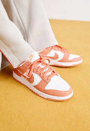 Persona che indossa sneakers Nike bianche e arancione chiaro con lacci, in piedi su un pavimento rivestito di moquette gialla con pantaloni beige.