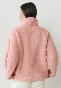 Giacca in pile rosa soffice con colletto alto, caratterizzata da una texture morbida e una vestibilità comoda. La parte posteriore presenta cuciture semplici e nessun ornamento.